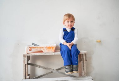Cute kid construction worker sitting on wooden table against white wall in apartment under renovation. Cheerful little boy wearing safety helmet and work overalls while playing at home.