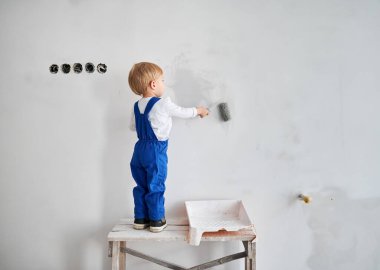 Ahşap masanın üzerinde duran ve apartmanın beyaz duvarını boyayan çocuk inşaat işçisinin arka görüntüsü. İş yerindeki bir çocuk, evde tadilat yaparken boya silindiri kullanıyor..