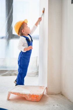Çocuk inşaat işçisi oturma odasında boya fırçasıyla duvar boyuyor. Güvenlik kaskı takan ve boya fırçası kullanan bir çocuk apartman tadilatında çalışıyor..