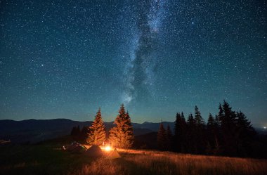 Yıldızlı gökyüzünün altındaki dağlarda gece kampı. Samanyolu ile dolu güzel gökyüzünün altındaki kamp alanında turist çadırları. Yürüyüşçülerin silueti yanan kamp ateşinin yanında dinleniyor. Turizm kavramı.