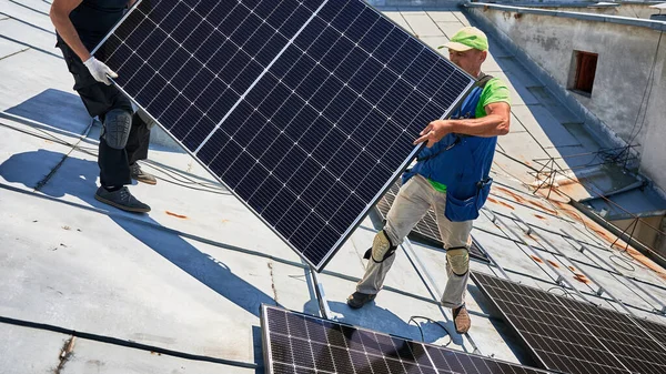 Workers building solar panel system on metal rooftop of house. Two men installers carrying photovoltaic solar module outdoors. Alternative, green and renewable energy generation concept.