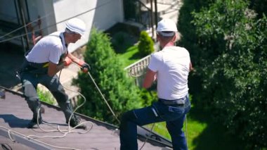 Erkek işçiler evin çatısına güneş paneli kuruyor. Kasklı elektrikçiler fotovoltaik güneş modülünü açık havada halatların yardımıyla kaldırıyorlar. Alternatif ve yenilenebilir enerji kavramı.