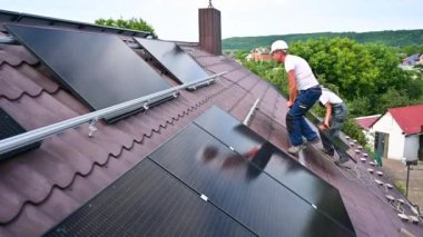 İşçiler evin çatısına güneş paneli inşa ediyorlar. Dışarıda fotovoltaik güneş modülü taşıyan kasklı iki adam var. Alternatif, yeşil ve yenilenebilir enerji üretimi kavramı.