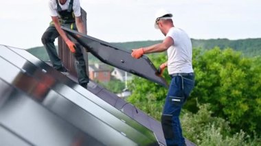 İşçiler evin çatısına güneş paneli inşa ediyorlar. Dışarıda fotovoltaik güneş modülü taşıyan kasklı iki adam var. Alternatif, yeşil ve yenilenebilir enerji üretimi kavramı.