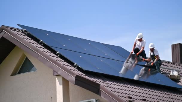 家の屋上にソーラーパネルシステムを構築する労働者 屋外に太陽光発電モジュールを設置するヘルメットに2人の男性インストーラー オルタナティブ グリーン 再生可能エネルギーのコンセプト — ストック動画
