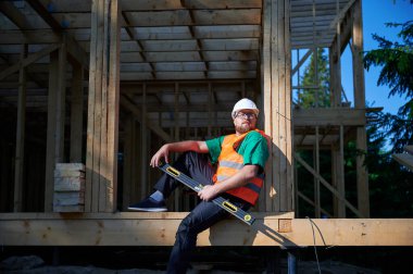 Marangoz ahşap çerçeve iki katlı ev inşa ediyor. Sakallı, gözlüklü, gözlüklü koruyucu kask takan, tulumlu ve turuncu yelek giyen bir adam. Modern ekolojik yapı kavramı.