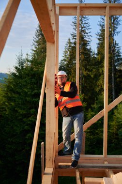 Marangoz ahşap çerçeve inşa ediyor. Gözlük takan sakallı adam, ruh seviyesini kullanarak duvarları kontrol ediyor. Çevre dostu çağdaş yapı kavramı.