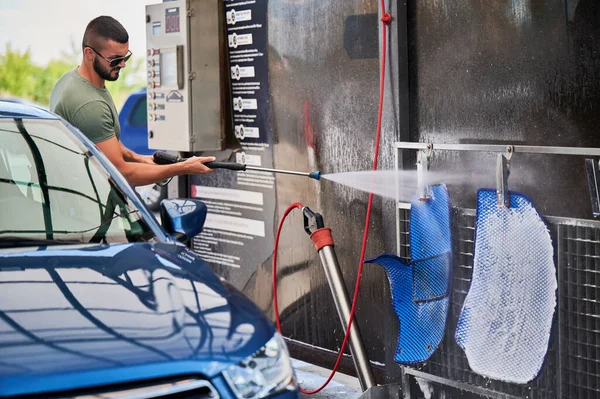 Confident man washing car mats on self-service car-washing station. Full car cleaning. Man in sunglasses cleaning blue mats from his auto using active foam.