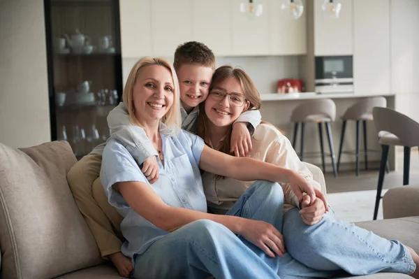 Lovely family relaxing on couch in living room. Little boy hugging his sister and mom. Cozy spending time with family indoors. In background, modern kitchen, neutral colors, minimalistic design.