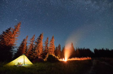Gece, yıldızlarla dolu dağların altında kamp kuruyor. Ormanın yakınındaki tepelerdeki aydınlık turist çadırlarının üzerinde yıldızlı gökyüzü. Gecenin karanlığında kamp ateşinden gelen sıcak ışık.