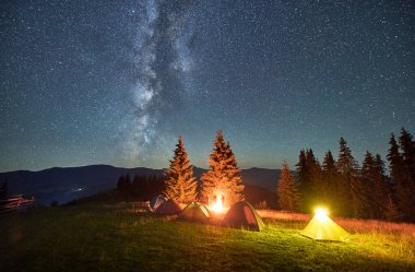 Yıldızlı gökyüzünün altındaki dağlarda gece kampı. Kamp alanındaki çimlerin üzerinde duran çadırlar, parlak ateş, gökyüzünde parlayan yıldızlar. Turizm ve macera kavramı.
