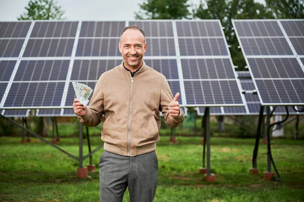 Successful investor happy about his profit. Male adult showing thumbs up and smiling straight to camera on background of solar panels.
