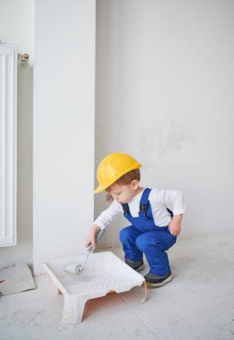 Küçük inşaat işçisi çocuk çömelmiş ve duvar boyası için boya silindiri hazırlıyor. Çocuk güvenlik kaskı takıyor ve evde tadilat yaparken iş tulumu giyiyor..