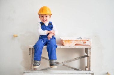 Şirin bir inşaat işçisi, tadilat altındaki beyaz duvarın karşısında, tahta masanın üzerinde oturuyor. Güvenlik kaskı takan ve kameraya bakıp gülen neşeli küçük çocuk..