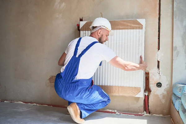 Male central heating technician crouching down and mounting heating radiator on the wall. Man wearing safety construction helmet and work overalls while working on home renovation.