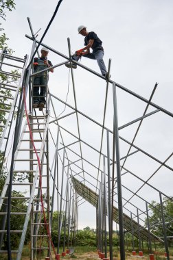 Güneş modülleri için metal demiryolu döşeyen güvenlik kasklı bir adam merdiven çıkıp çekiç tutuyor. Beyaz gökyüzü altında güneş panelleri için destek yapıları oluşturan erkek işçiler.