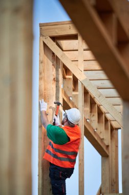 Marangoz ormanın yakınında ahşap çerçeve inşa ediyor. Adam koruyucu kask ve inşaat yeleği giyerken çivi çakıyor. Modern ekolojik yapı kavramı.