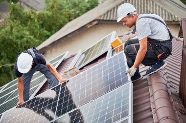 Erkek teknisyenler fotovoltaik güneş modüllerini evin çatısına monte ediyorlar. Kasklı elektrikçiler dışarıda güneş paneli sistemi kuruyorlar. Alternatif ve yenilenebilir enerji kavramı.
