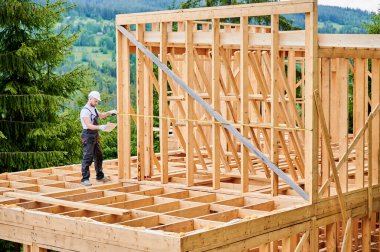 Marangoz ahşap iskelet evi inşa ediyor. Adam iş kıyafeti ve güvenlik kaskı giyerken mesafeyi mezurayla ölçüyor. Çağdaş, çevre dostu inşa uygulamalarının ilkeleri.