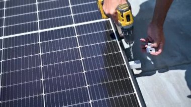 Metal çatı katında fotovoltaik güneş paneli inşa eden bir işçi. İnsan teknisyeninin güneş modülünü açık havada elektrikli matkapla kurmasına yardım edin. Alternatif enerji üretimi kavramı.