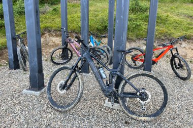 Dağlarda çakıl kaplı bir zemine park edilmiş dağ elektrikli bisikletleri koleksiyonu. MTB motorları kalın siyah sütunlu bir yapının altına yerleştiriliyor..