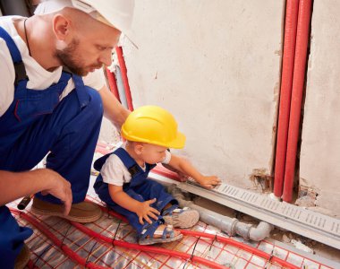 Çocuk borularla duvarda oturuyor ve erkek tesisatçıyla tesisat sistemi kuruyor. Baba ve oğul güvenlik miğferinde ev tadilatında birlikte çalışıyorlar..