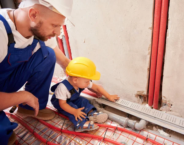 Çocuk borularla duvarda oturuyor ve erkek tesisatçıyla tesisat sistemi kuruyor. Baba ve oğul güvenlik miğferinde ev tadilatında birlikte çalışıyorlar..
