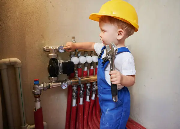 儿童在安全施工头盔持有扳手工具和指向温度计 同时检查家庭管道安装 维修进行中的公寓水管系统的小孩 图库图片