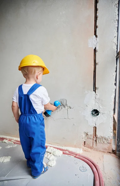 Back view of child construction worker drilling wall with toy electric drill tool in apartment. Kid wearing safety helmet and work overalls while working on home renovation.