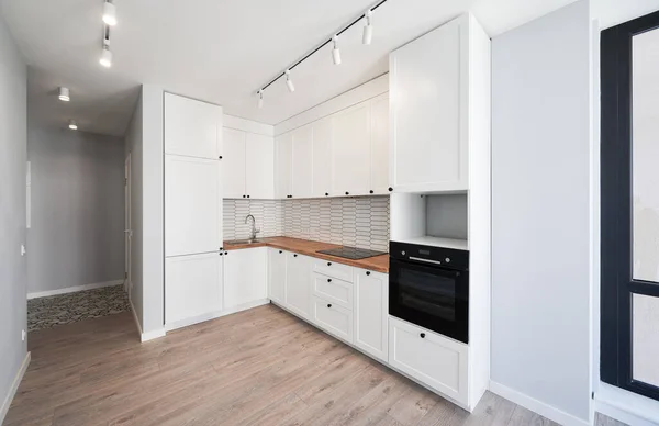 Kitchen with laminate floor, white walls, doorway and modern kitchen set with electric stove, counter, sink and oven. Apartment kitchen room with elegant minimalist design in white colors.
