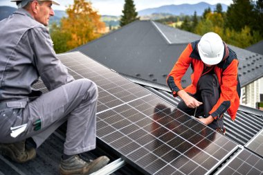 Mühendisler evin çatısına fotovoltaik güneş panelleri kuruyor. Miğferli adam teknisyenleri büyücülerin yardımıyla güneş modülü sistemi inşa ediyorlar. Alternatif, yenilenebilir enerji kavramı.