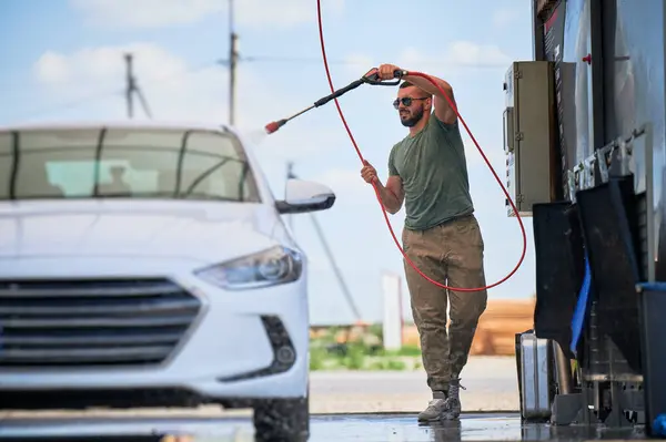 Washing luxury white auto with water gun on an open air car wash. Confident man cleaning his car with high pressure water jet. Technical service concept.
