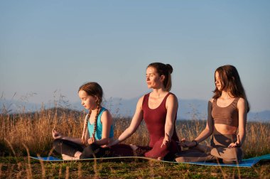 Genç anne kızlarıyla meditasyon yapıyor. Şafakta dağlarda meditasyon. Sabahın erken saatlerinde lotus pozisyonunda oturan üç kadın..