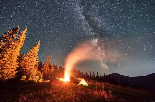 Night camping under sky full of stars and Milky way in the mountains. Starry sky over illuminated tourist tents on hills near forest. Warm light from campfire at dark night.