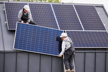 Elektrikçiler evin çatısına güneş paneli kuruyor. Miğferli işçiler fotovoltaik güneş modülünü dışarı halatların yardımıyla kaldırıyorlar. Alternatif ve yenilenebilir enerji kavramı.