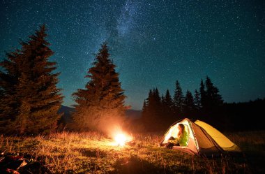 Night camping in mountains under starry sky and Milky way. Female tourist sitting in tent entrance in campsite, admiring landscape and burning campfire. Concept of traveling and hiking.