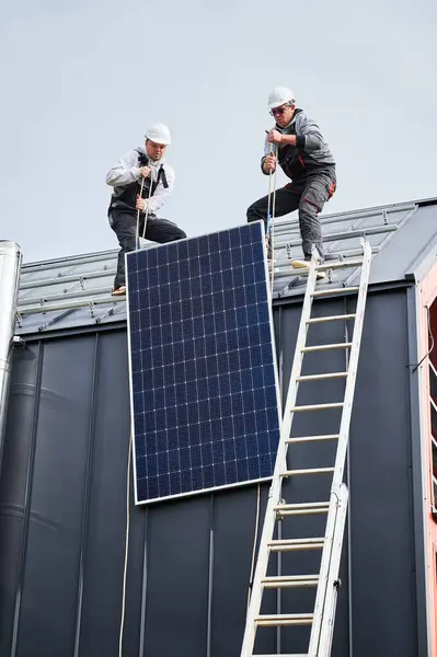 Men builders installing solar panel system on roof of house. Electricians in helmets lifting up photovoltaic solar module with help of ropes outdoors. Concept of alternative and renewable energy.