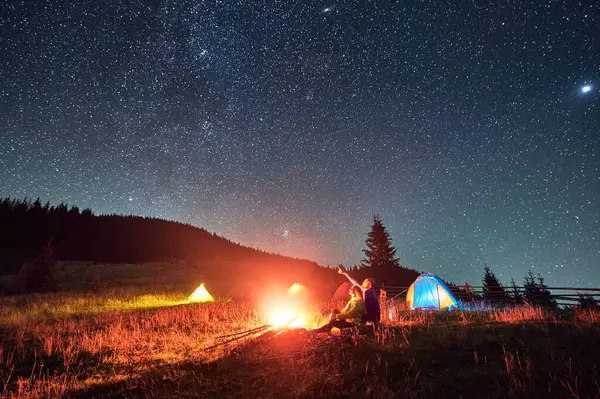 Night camping in mountains under starry sky. Two people, couple sitting on grass near burning campfire. Man pointing on the beautiful sky full of stars. Concept of tourism and healthy living.