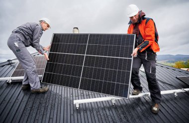 Evin çatısına güneş paneli inşa eden mühendisler. Dışarıda fotovoltaik güneş modülü taşıyan miğferli işçiler. Alternatif ve yenilenebilir enerji kavramı.