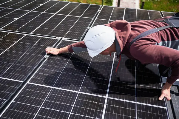 Man builder mounting photovoltaic solar panels on roof of house. Engineer in helmet installing solar module system with help of hex key. Concept of alternative, renewable energy.