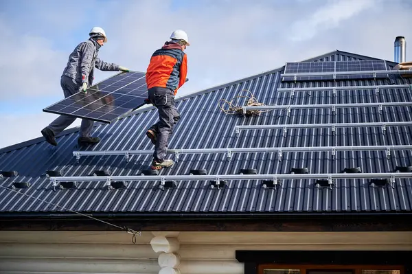 屋顶上建造太阳能电池板系统的屋顶 戴钢盔的男工在室外携带光电太阳能组件 替代能源和可再生能源的概念 免版税图库图片