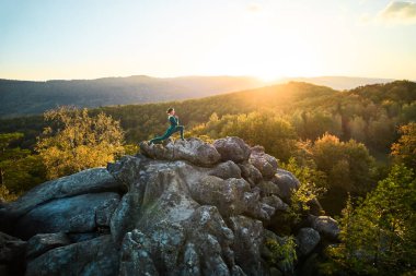 Yoga ve meditasyon yapan bir kadın akşamları dev bir kayanın tepesinde. Güzel günbatımı, sonbahar ormanları, arka planda kayalar ve tepeler. Hava aracı görünümü