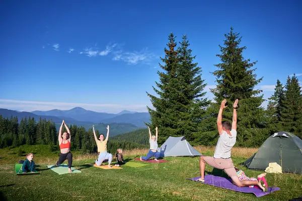 Group of people doing yoga pose outdoor in camping in the mountains. Adults and children standing on yoga mats, each doing yoga pose under clear blue sky in morning.