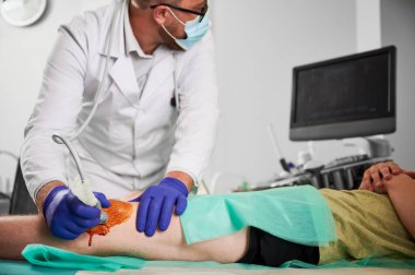 Ofisinde diz çöküp hasta muayene eden erkek uzman, otobüste yatan erkek hasta. Beyaz bornoz, lateks eldiven, gözlük ve maske takan bir doktor. Kontrol ve sağlık hizmetleri kavramı.