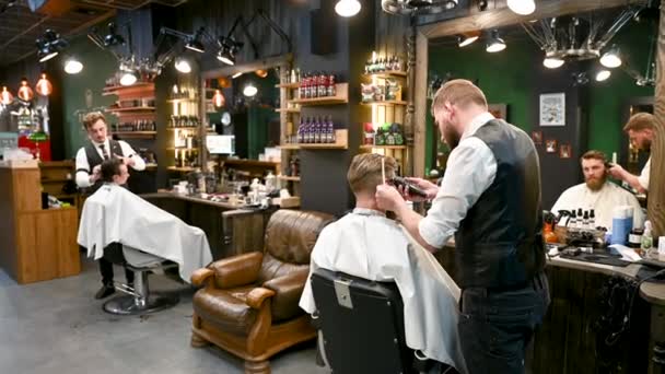 側面の毛を切るのに櫛および電気シェーバーを使用する専門の棒 スタイリッシュなヘアカットを得る男性の顧客 他の美容師の背景に男性の側面のビュー — ストック動画