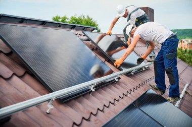 İşçiler evin çatısına güneş paneli inşa ediyorlar. Dışarıda fotovoltaik güneş modülü taşıyan kasklı iki adam var. Alternatif, yeşil ve yenilenebilir enerji üretimi kavramı.