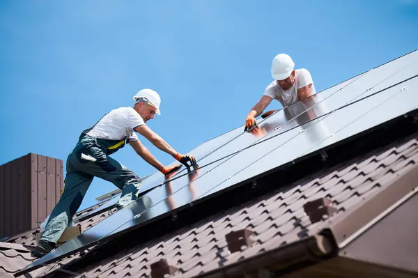 stock image Workers building solar panel system on rooftop of house. Two men installers in helmets installing photovoltaic solar module outdoors. Alternative, green and renewable energy generation concept.