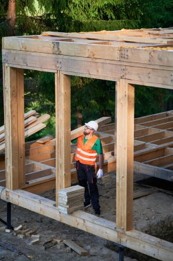 Marangoz ormanın yakınında ahşap çerçeve inşa eder. Adam omuzunda büyük bir kiriş tutuyor, iş kıyafetleri ve kask giyiyor. Modern ekolojik yapı kavramı.