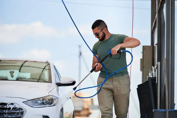 Washing luxury white auto with water gun on an open air car wash. Confident man cleaning his car with high pressure water jet. Technical service concept.