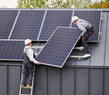 Bağlayıcılar evin çatısına güneş paneli kuruyor. Dışarıda fotovoltaik güneş modülü taşıyan miğferli işçiler. Alternatif ve yenilenebilir enerji kavramı.